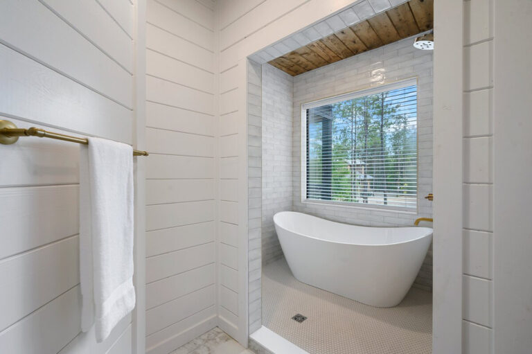Freestanding soaking tub with brass fixtures positioned beneath a large picture window in a serene bathroom setting.