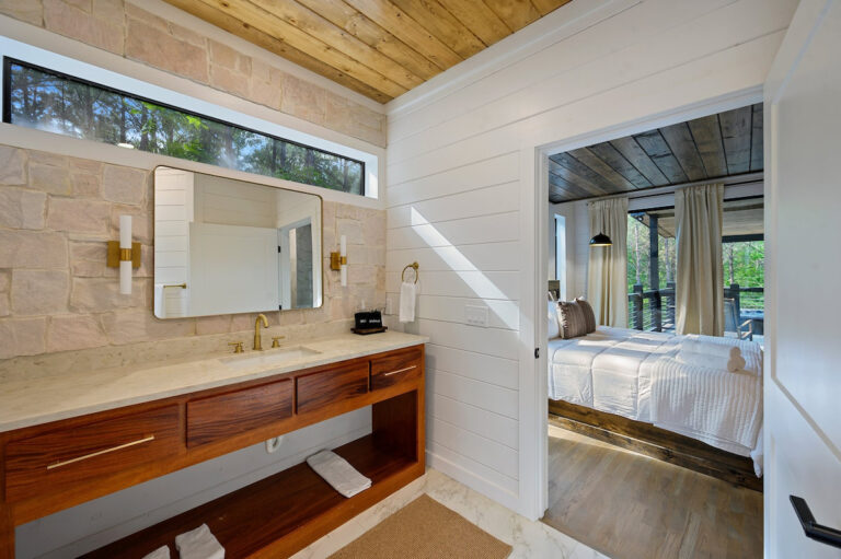 Bathroom vanity with a wide mirror and window, reflecting the bedroom visible through the open doorway.