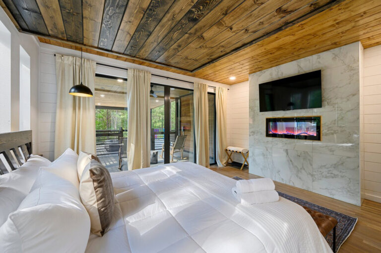 Modern bedroom with fresh white bedding, built-in wall fireplace, and large picture windows providing natural light.