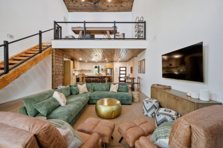 Living room with cozy seating and a stone fireplace, with a modern kitchen visible in the background.