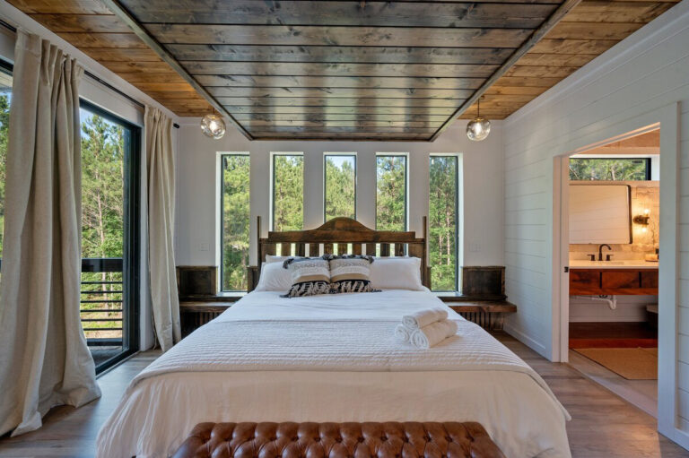 Rustic-style bedroom with white bedding and large picture windows letting in sunlight and forest views.