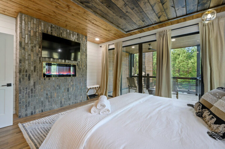 Bedroom with a cozy bed, warm-toned bedding, and large picture windows offering views of the surrounding trees.