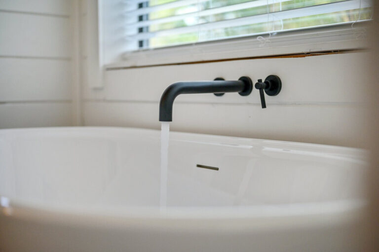 Close-up of a soaking tub with matte black faucet and controls, large window in background.