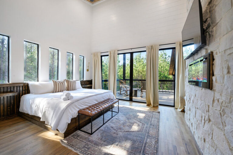 Modern bedroom featuring white linens, wood-accented décor, and tall windows that brighten the room.