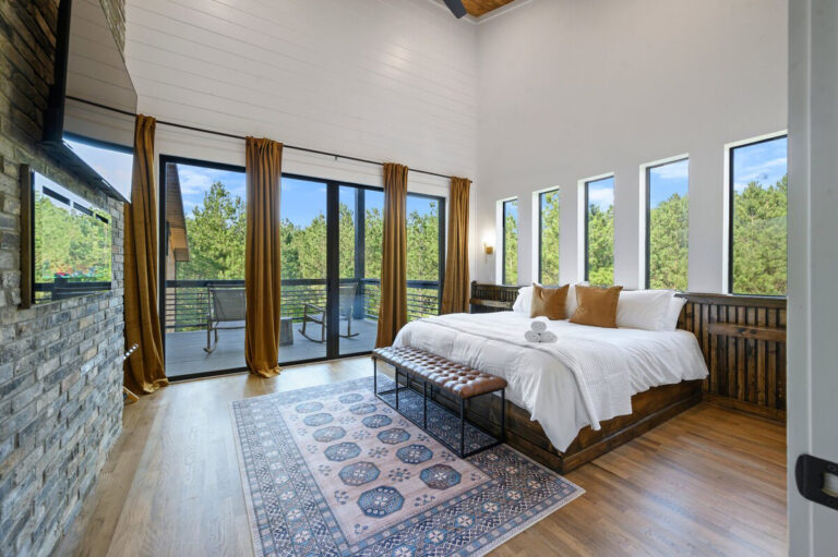 Cozy bedroom with a large bed dressed in white bedding and warm brown accents, illuminated by natural light from expansive picture windows.