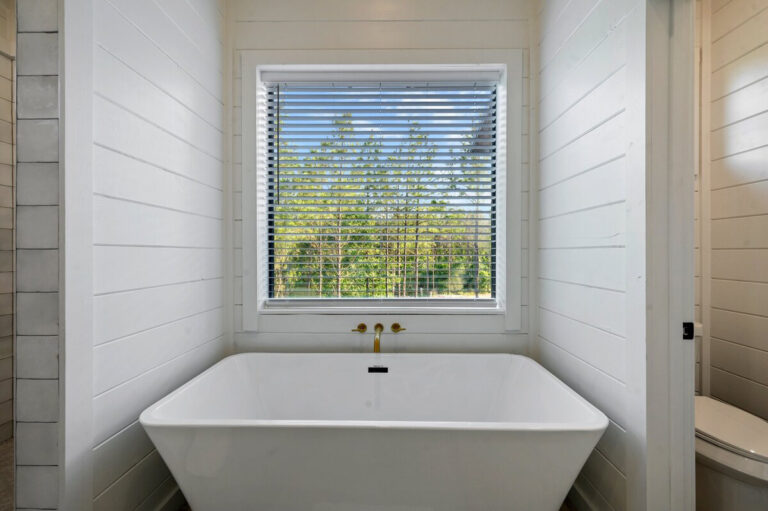 Spacious modern soaking tub with brass fixtures, surrounded by white paneling and positioned beneath a large picture window.