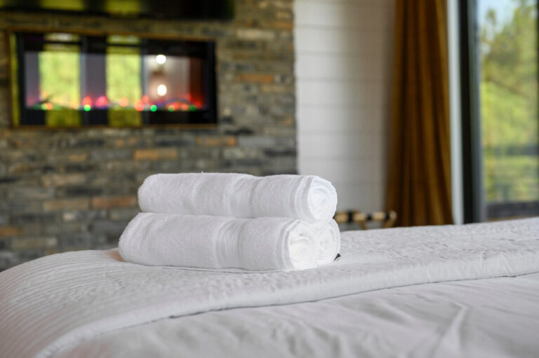 Rolled towels resting on a textured throw blanket at the foot of a wood-framed bed with a fireplace in the background.