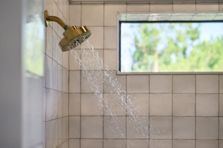 Close-up of a brass shower head mounted against white tile with a nearby window providing natural light.