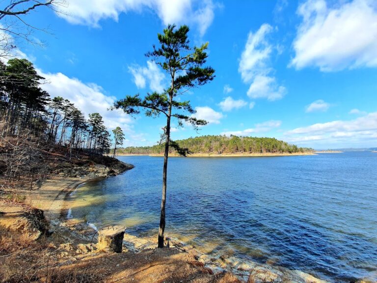 A serene view of the lake at beavers bend state park, with calm, crystal-clear water reflecting the surrounding forested hills under a bright blue sky.