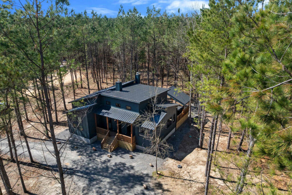 Aerial view of blue note broken bow cabin surrounded by pine trees near outdoor fire pit and river activities.