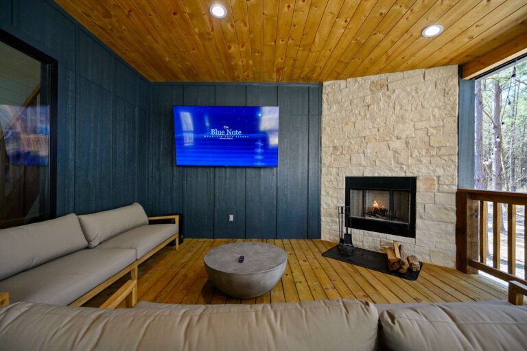 Covered outdoor lounge at Blue Note Broken Bow cabin with fireplace, TV, and comfortable seating overlooking wooded scenery.