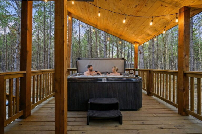 Couple relaxing in hot tub at Blue Note Broken Bow cabin beneath string lights in peaceful wooded outdoor retreat.