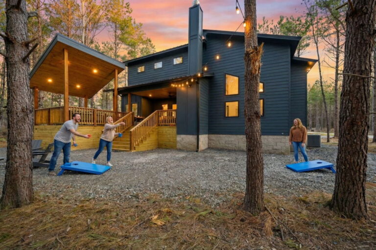 Guests playing cornhole outside Blue Note Broken Bow luxury cabin with covered deck, string lights, and wooded retreat setting.