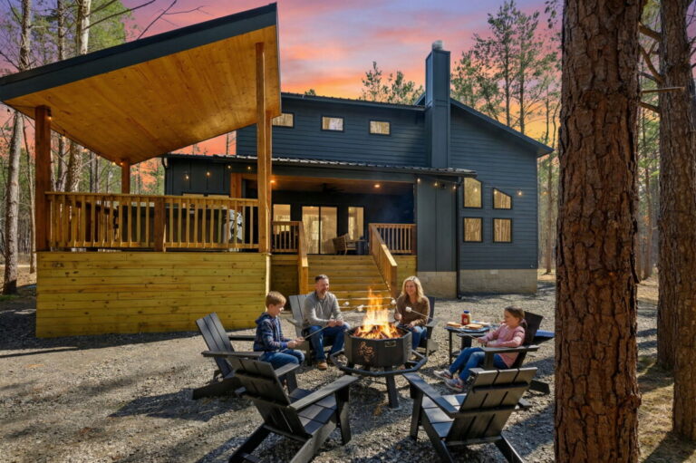 Family roasting marshmallows around outdoor fire pit at Blue Note Broken Bow cabin during sunset in wooded retreat setting.